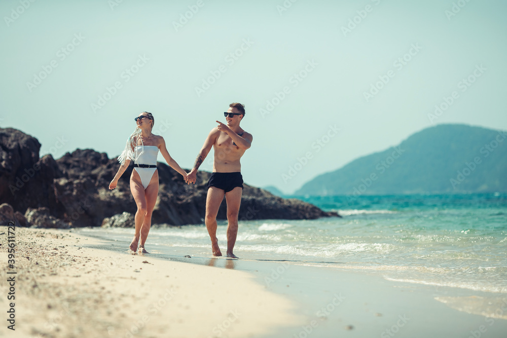 Outdoor shot of romantic hipster young couple walking along the sea shore holding hands. Young man with tattoo and woman with white dreadlocks walking on the beach together. Beautiful sea. Thailand