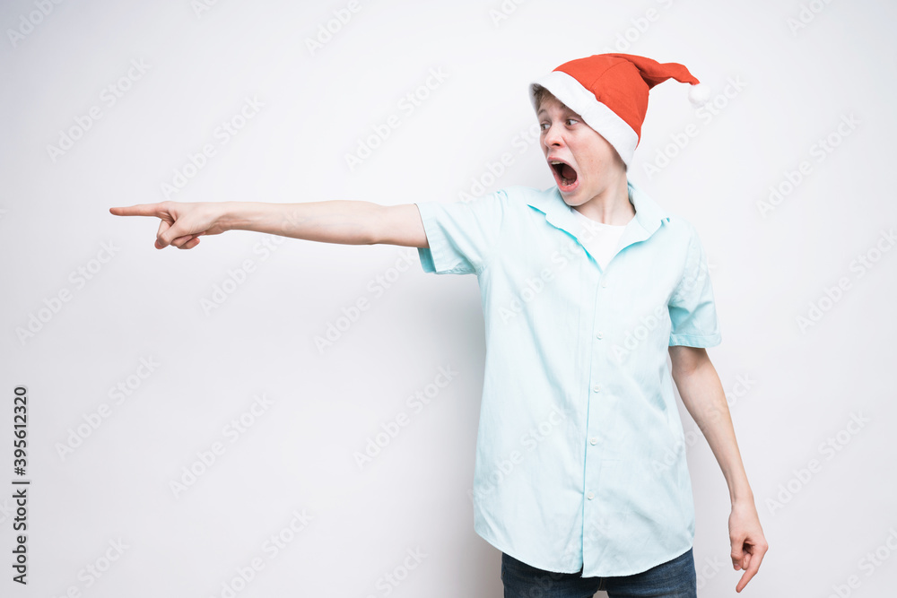 A caucasian boy in Santa Claus hat is pointing to something on a white background