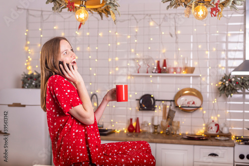 Happy laughing and smiling young beautiful woman speaking by mobile phone in kitchen in red pygamas holding tea mug. New year and xmas alone at home photo