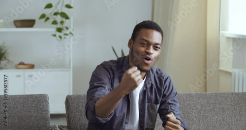 Happy black man young guy fan cheerleader sitting on couch watching sports program on TV rejoices winning victory of adored favorite team makes gesture of admiration delight triumph applauds says yes