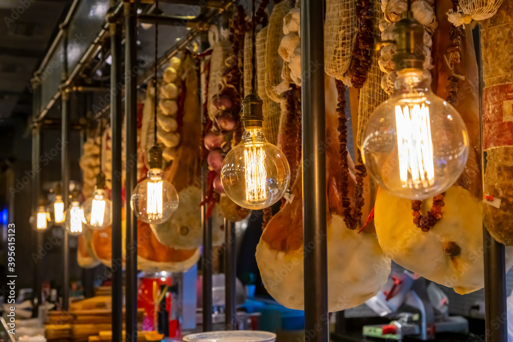 Prosciutto (dry-cured ham) legs meat, different sausages and a bunch of garlic and pepper hangs in the restaurant ready for sale. Incandescent lamps hangs nearby. Dry-cured meat theme.