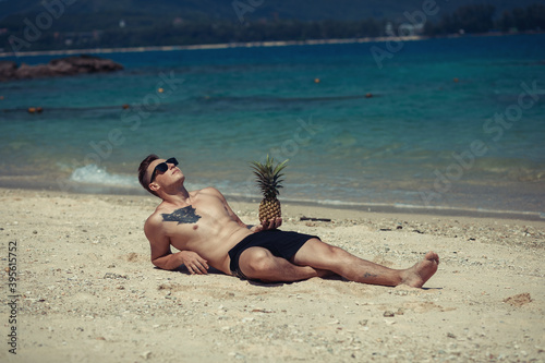happy shirtless young hipster man  in shorts and sunglasses  with tattoo lying and holding pineapple on sea beach. Summer vacation. Tropical seascape with boats on the background.