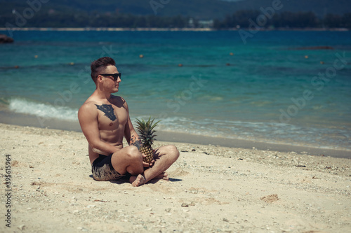happy shirtless young hipster man  in shorts and sunglasses  with tattoo lying and holding pineapple on sea beach. Summer vacation. Tropical seascape with boats on the background..