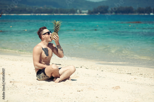 Stylish muscular hipster young man with tattoo wearing sunglasses trying to eat pineapple and sitting on the beach. Summer time concept. Phuket. Thailand