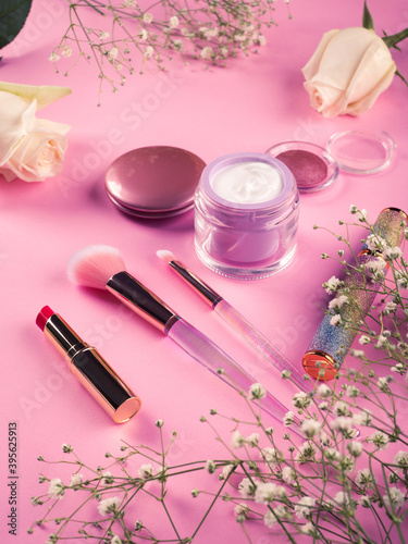 Shiny make up products and accessories on pink background with flowers. Trendy holographic brushes.