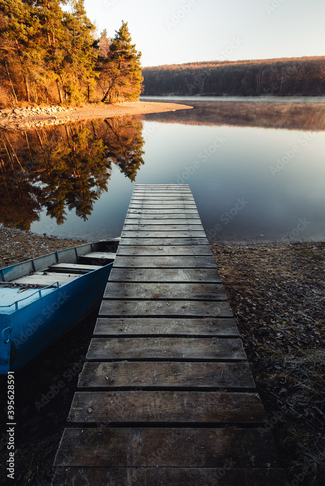 Fototapeta premium Moody mole (pier) and old blue boat in fall with crystal blue water and forest on background. Sunrise - mole (jetty) on the lake with water reflection of forest.