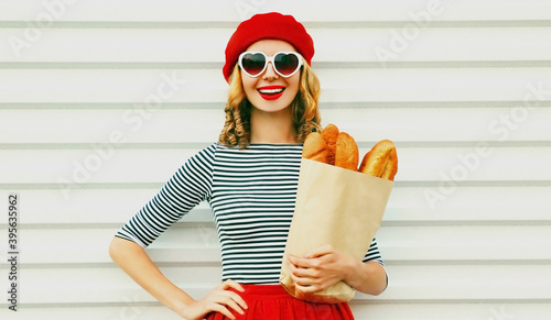 Close up of laughing young woman wearing a french red beret holding paper bag with long white bread baguette over a white background photo