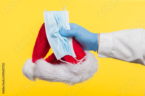Close-up profile side view of a human hand in blue surgical gloves, holding a Santa Claus hat and a protective mask against COVID-19 coronavirus infections, isolated on yellow background.
