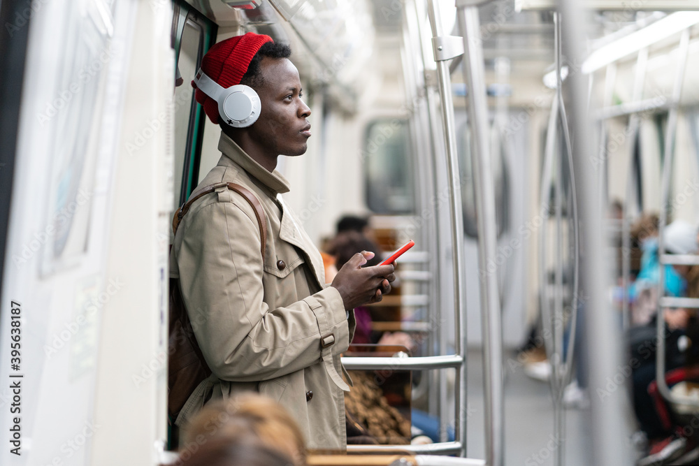 Afro-American passenger man in red hat, trench coat stand in subway train, using mobile smart phone, listens to music with wireless headphones in public transportation.  