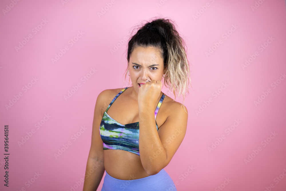Naklejka premium Young beautiful woman wearing sportswear over isolated pink background angry and mad raising fist frustrated and furious while shouting with anger. Rage and aggressive concept.