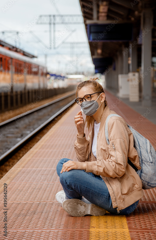 Woman in a mask on the platform near the train. The girl is sitting at the train station near the train. Travel by rail. View of a traveler during an epidemic. Travel and Covid-19. Bydgoszcz.