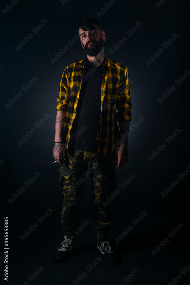 Full-length portrait of an attractive man with tattoos posing dressed in yellow and black checkered shirt on a black background