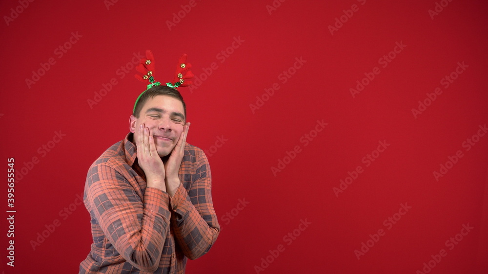 A young man stands with a headband in the form of Christmas horns and is shy. Studio photography on a red background.