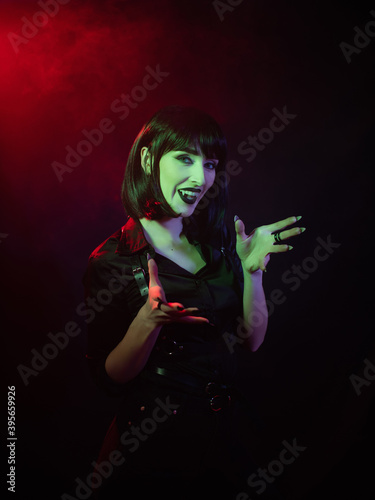 a girl in the guise of a vampire in a green-red light and smoke spread her arms and looks into the camera, with her mouth open where vampire fangs are visible
