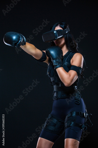 A young athletic woman in an EMS suit and virtual reality glasses with Boxing gloves on an isolated black background. EMS training. Electrical muscle stimulator.
