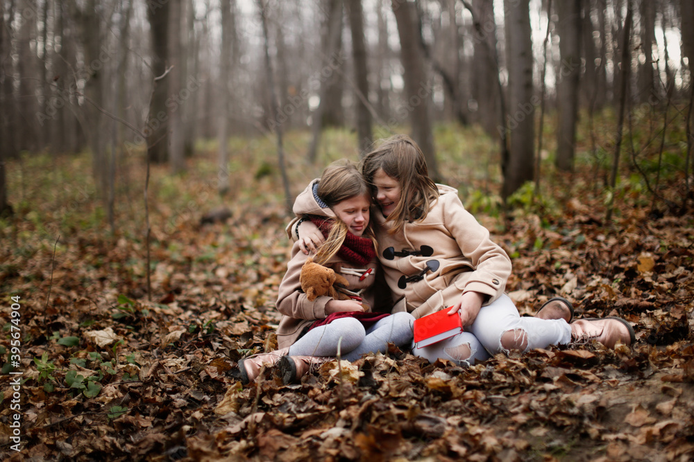 Caucasian children sisters hugs in park read a book on a picnic, children in the forest sit with a book in a meadow, autumn walks in warm coats Caucasian children sisters hugs in park read a book on a picnic, children in the forest sit with a book in a meadow, autumn walks in warm coats