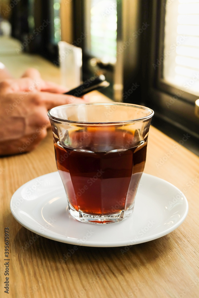 Cup of drip americano coffee with white saucer on wooden table against blurred male hands holding smartphone background. Cup of drip americano coffee with white saucer on wooden table against blurred male hands holding smartphone background.