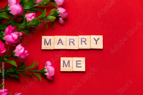 Wooden block lettering "marry me" on red textured background with bouquet of roses: wedding background, top view