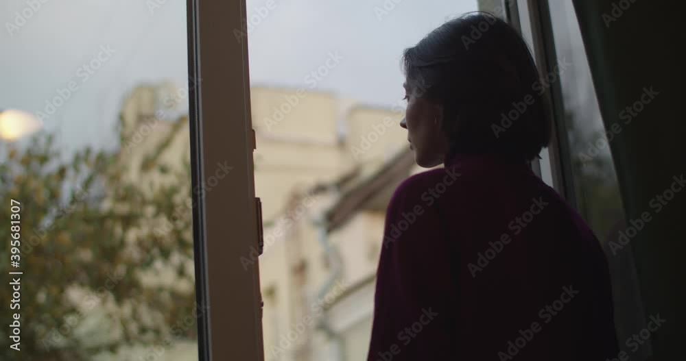 Back low angle view of girl standing by open window breathing slow motion. Young woman looking outdoors with thoughtful expression silhouetted in evening light. Depression loneliness quarantine Back low angle view of girl standing by open window breathing slow motion. Young woman looking outdoors with thoughtful expression silhouetted in evening light. Depression loneliness quarantine