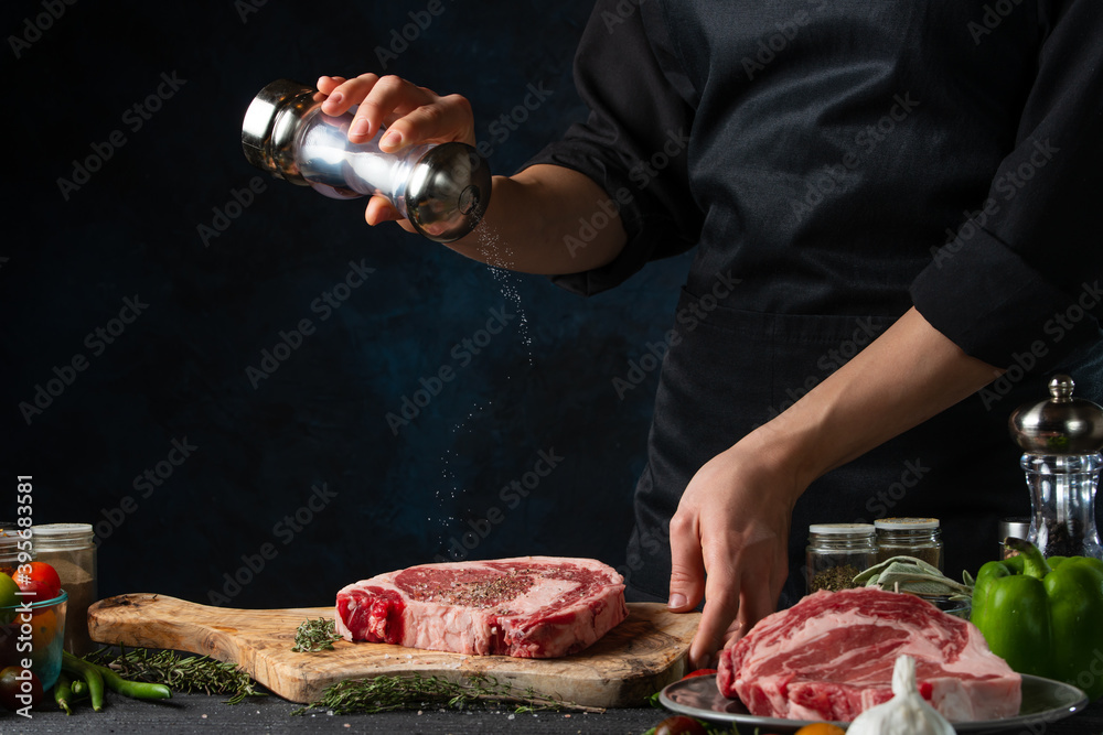 Close-up view of chef salts raw steak on wooden chopped board. Backstage of preparing grilled pork meat at restaurant kitchen on dark blue background. Frozen motion. Cooking process.