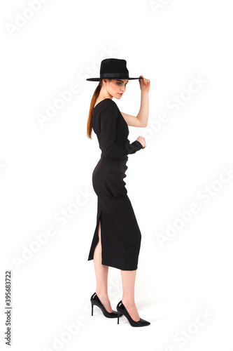 Stylish young businesswoman in black elegant dress and hat on white isolated background. Woman in fashionable clothes posing at camera. Concept of style, fashion, beauty and achievement of goals