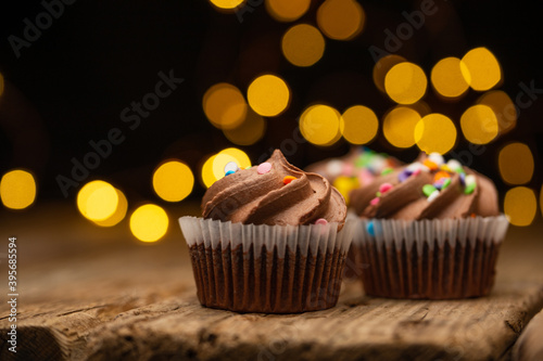 Tasty chocolate cupcakes with sprinkling on rustic wooden table on black background with yellow lights. Sweet dessert. Bakery concept. Elegant food. Sweets for coffee or tea. Space for text. Close-up