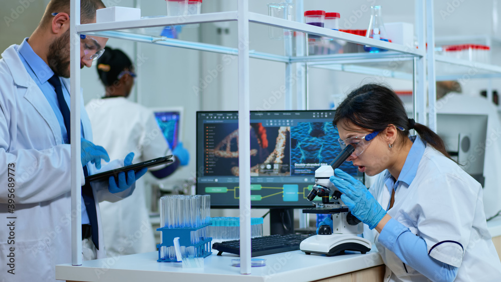 Scientist team checking virus development using microscope and writing analysis results on tablet in modern equipped lab. Multiethnic group examining vaccine evolution using high tech for research. Scientist team checking virus development using microscope and writing analysis results on tablet in modern equipped lab. Multiethnic group examining vaccine evolution using high tech for research.