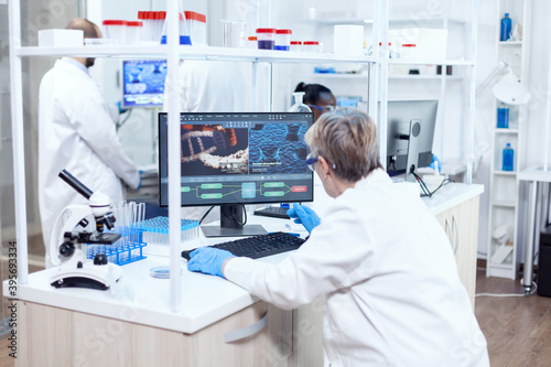 Senior scientist in pharmaceuticals laboratory doing genetic research wearing lab coat with team in the background. Male chemist engineering vaccine working on computer.