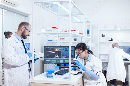 Male scientist taking notes on tablet pc during virus study. Team of researchers doing pharmacology engineering in sterile laboratory for healthcare industry with african assistant in the background.