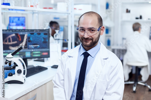 Pharmaceutical researcher sitting on stool looking at camera.Serious expert in genetics in lab with modern technology for medical investigations with african assistant in the background.
