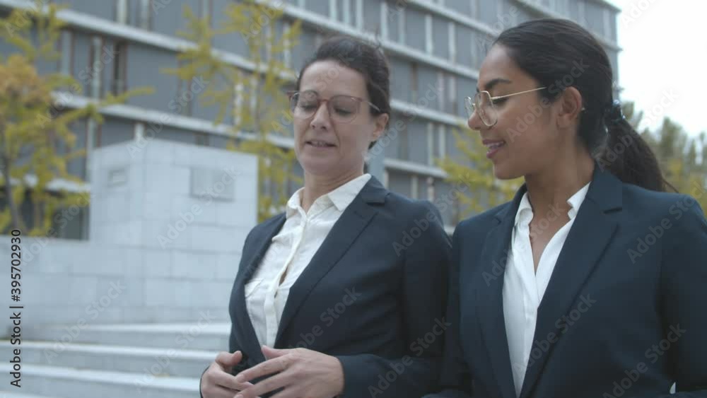 Two female office friends walking near office building and talking, chatting outside, discussing work, colleagues, career or news. Dolly shot. Business communication concept. Two female office friends walking near office building and talking, chatting outside, discussing work, colleagues, career or news. Dolly shot. Business communication concept.