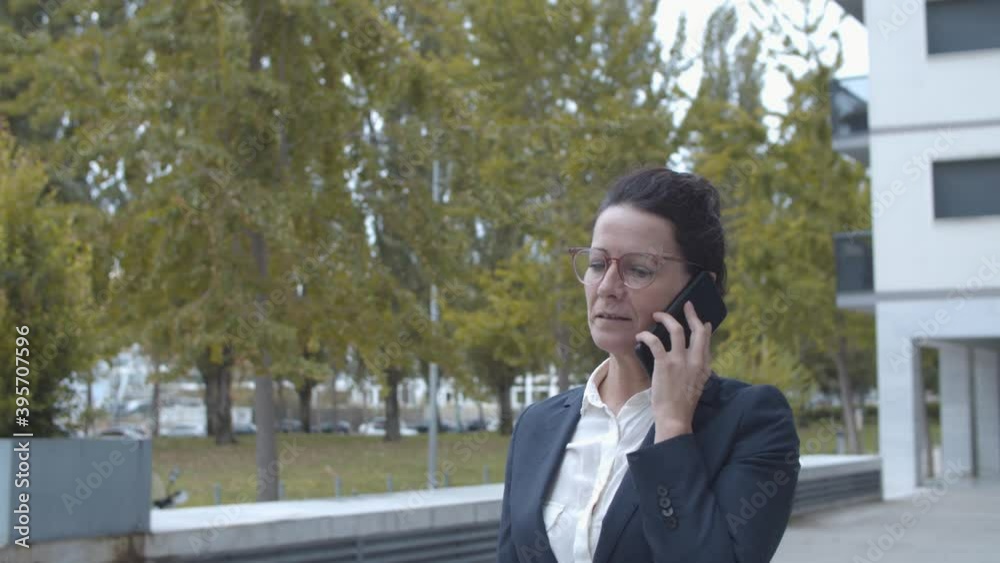 Happy confident businesswoman in office suit talking on cellphone while walking outside, speaking, smiling and making winners gesture. Dolly shot. Business communication concept. Happy confident businesswoman in office suit talking on cellphone while walking outside, speaking, smiling and making winners gesture. Dolly shot. Business communication concept.