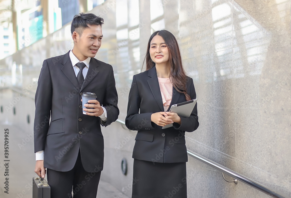young Asian business man holding cup of coffee walking in public walkway with young business woman , smiling and talking together. young Asian business man holding cup of coffee walking in public walkway with young business woman , smiling and talking together.