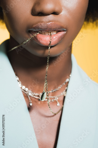 cropped view of african american woman in blue stylish outfit with necklace in mouth isolated on yellow © LIGHTFIELD STUDIOS