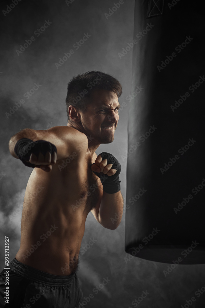 Aggressive boxer in black boxing wraps punching in boxing bag on dark background with smoke