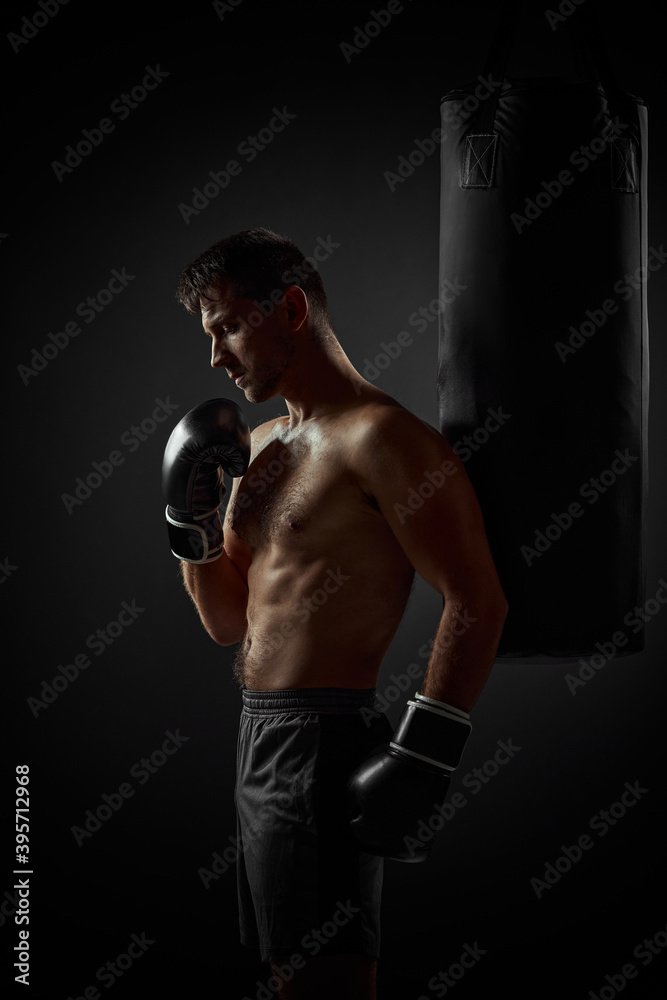 Young handsome male boxer in black gloves standing near punching bag on dark background