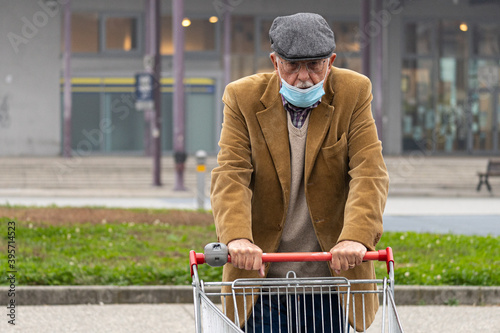 Ederly man wearing coronavirus protective surgical mask with shopping cart. Pensioner ready for shopping, as fear and anxiety for shortages during COVID-19 pandemic spred photo