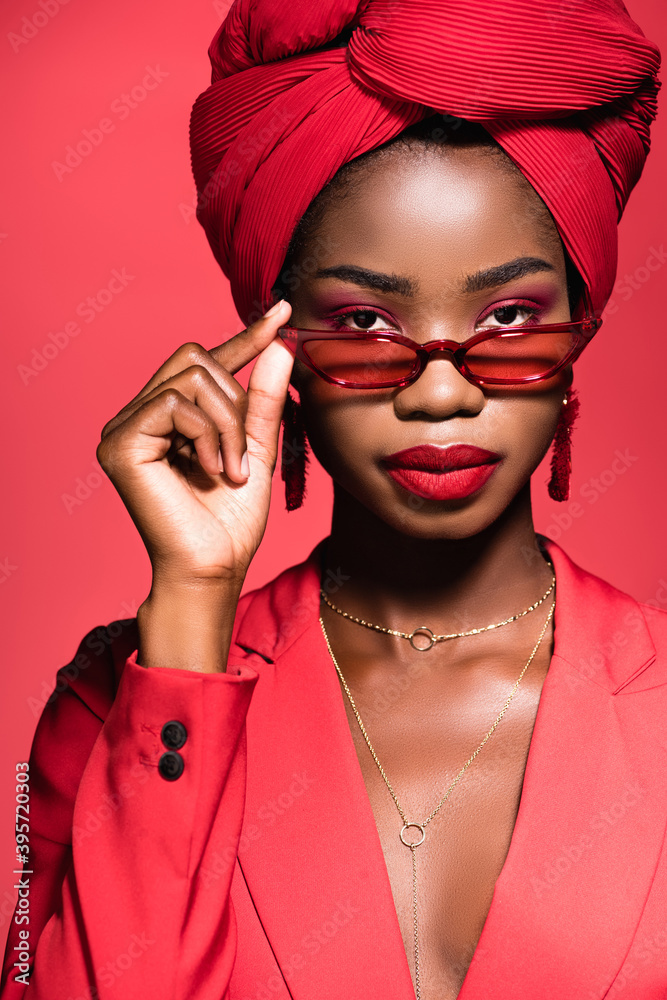 Φωτογραφία african american woman in stylish outfit, sunglasses and turban isolated on red