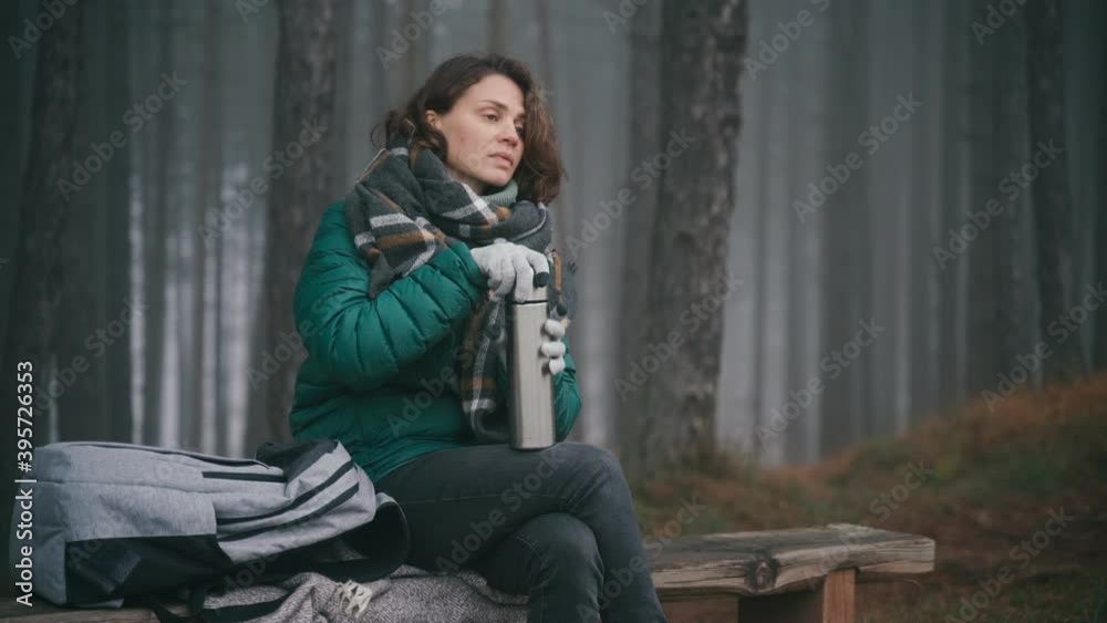 Young woman taking a thermos out of her backpack and drinks a hot tea while sitting on a wooden bench in a misty autumn forest. Young woman taking a thermos out of her backpack and drinks a hot tea while sitting on a wooden bench in a misty autumn forest.