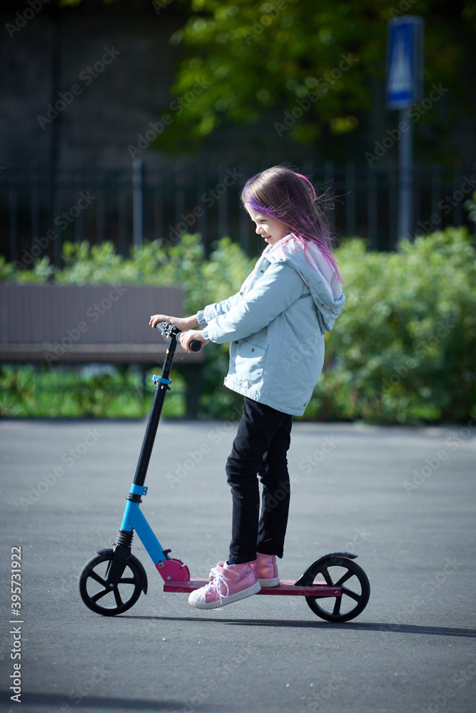 Smiling little girl rides scooter in park. Empty space for text