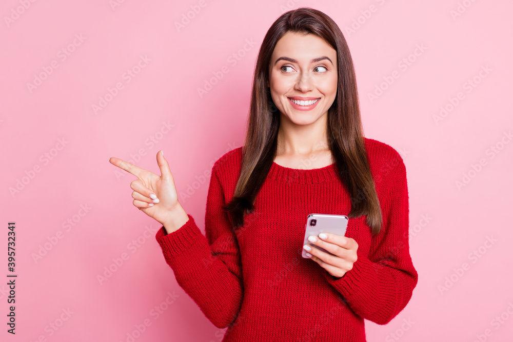 Photo of lovely girl directing forefinger look side empty space hold cellphone shiny smile wear red sweater isolated pink color background Photo of lovely girl directing forefinger look side empty space hold cellphone shiny smile wear red sweater isolated pink color background