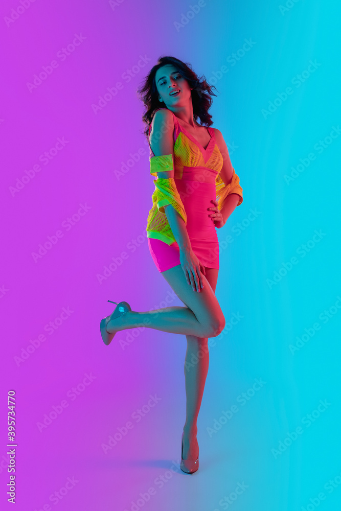 Dancing. Beautiful seductive girl in fashionable bright crimson and yellow dress on gradient pink-blue neon background. Full-length portrait. Copyspace for ad. Summer, fashion, emotions concept.