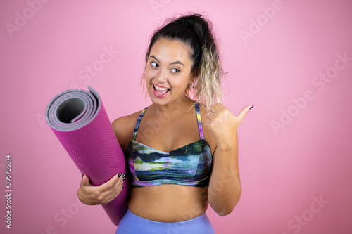 Young beautiful woman wearing sportswear and holding a splinter over isolated pink background pointing thumb up to the side surprised
