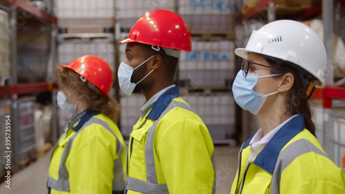 Side view diverse team of industrial workers in helmet and safety mask walking in large warehouse