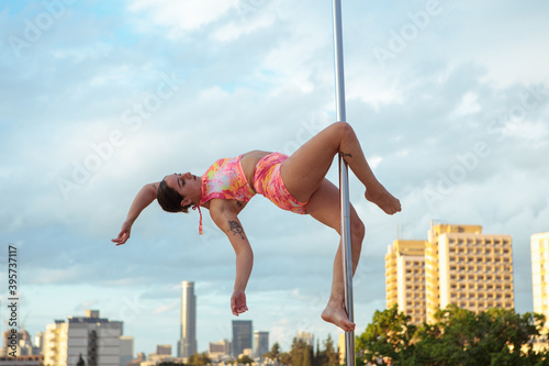 Young Middle Eastern pole dancer, holding a pose on a pole set outdoors. Tel Aviv buildings in the far background.