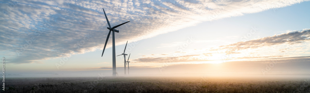 Three commercial wind turbines in thick fog at sunrise in the English countryside panoramic