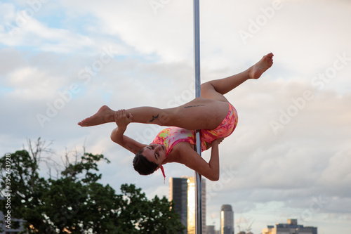 Young Middle Eastern pole dancer, holding a pose on a pole set outdoors. Tel Aviv buildings in the far background.
