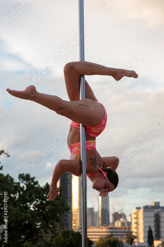 Young Middle Eastern pole dancer, holding a pose on a pole set outdoors. Tel Aviv buildings in the far background.
