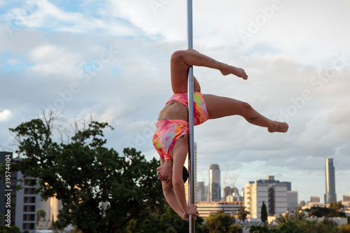 Young Middle Eastern pole dancer, holding a pose on a pole set outdoors. Tel Aviv buildings in the far background.