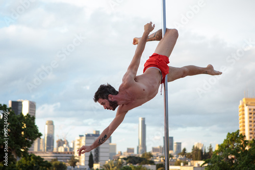 Muscular male pole dancer, holding a pose on a pole set outdoors. Tel Aviv buildings in the far background.